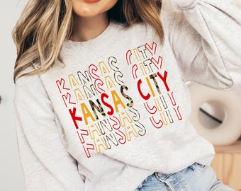 May include: A gray sweatshirt with the words "Kansas City" repeated in a stacked pattern. The text is in red, yellow, and black with a leopard print pattern.