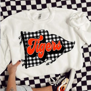 May include: White sweatshirt with a black and white checked flag design and the word "Tigers" in orange and black. This casual sweatshirt is made from a soft material and is suitable for everyday wear.