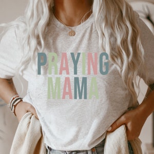 May include: A white t-shirt with the text "Praying Mama" in colorful letters. The letters are in shades of green, pink, and blue.