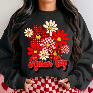 May include: Black sweatshirt with a floral design. The flowers are red, white, and checkered, each with a yellow smiley face. The words "Kansas City" are in red script across the bottom.