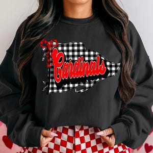 May include: Black sweatshirt featuring a buffalo plaid design with the word "Cardinals" in red script. A red bow is tied to the top left of the design. The sweatshirt is paired with red and white checkered pants.