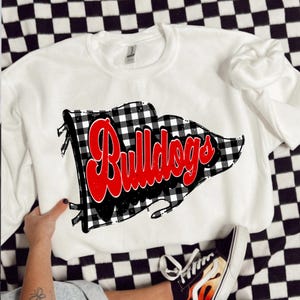 May include: White crew neck sweatshirt with a black and white buffalo check flag design and the word "Bulldogs" in red script. The sweatshirt is displayed on a black and white checkered surface.