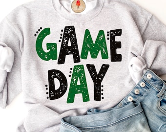May include: A gray sweatshirt with the words "GAME DAY" printed in black and green letters with a distressed look. The sweatshirt is folded and lying on a pair of blue jeans.