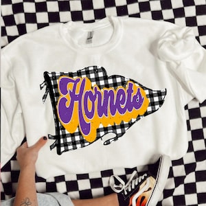 May include: White sweatshirt with a graphic reading "Hornets" in purple and yellow, set against a black and white checkered flag design. The sweatshirt is shown on a checkered surface.