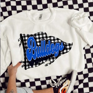 May include: White crew neck sweatshirt featuring a black and white buffalo check flag design. The word "Bulldogs" is written in blue script across the flag. The sweatshirt is displayed on a black and white checkered surface.