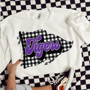 May include: White sweatshirt featuring a black and white checked flag design with the word "Tigers" in purple script. The sweatshirt is shown on a black and white checked surface.