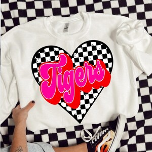 May include: White sweatshirt with a black and white checkered heart design. The word "Tigers" is written in pink and red letters inside the heart.