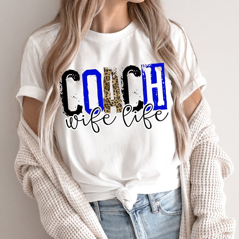 Może przedstawiać: Biała koszulka z czarnym i niebieskim tekstem, kt&oacute;ry brzmi "COACH wife life" z wzorem lamparta w środku słowa "COACH".