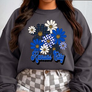 May include: Dark grey sweatshirt with a floral design in blue, white, and navy, including a checkerboard pattern. The words "Kansas City" are written in a retro font in blue and white.