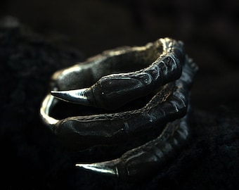 May include: A dark, textured ring shaped like a claw. The ring features three sharp, silver-colored talons extending from a dark, metallic band. The claw design gives the ring a gothic or fantasy aesthetic.
