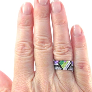 以下が含まれることがあります： 緑、白、紫、黒のパターンでビーズでできたリングをつけた手