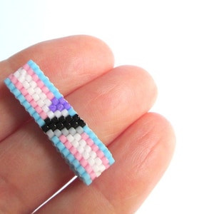 以下が含まれることがあります： トランスジェンダーのプライドフラッグの色、薄い青、ピンク、白、黒、薄い紫のビーズリングです。