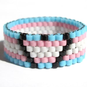 May include: A beaded ring with a pattern of blue, pink, white, and black beads. The beads are arranged in a pattern that resembles the transgender pride flag.