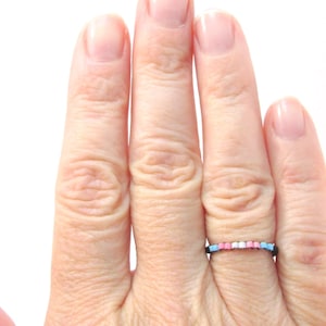 Könnte beinhalten: Eine Hand trägt einen Ring aus kleinen, bunten Perlen. Der Ring ist schwarz mit blauen, rosa und weißen Perlen.