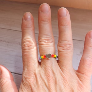 Könnte beinhalten: Ein Perlenring mit Regenbogenfarben und einem silbernen Herzanhänger. Der Ring besteht aus kleinen, farbigen Perlen in einem Farbspektrum, mit einem kleinen herzförmigen Anhänger in der Mitte.
