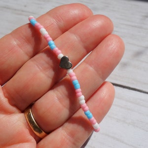 May include: A beaded bracelet featuring pink, white, and light blue beads. A small silver heart charm is centered on the bracelet. The bracelet is held up against a light wood background.