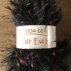 Pode incluir: Um novelo de lã preta Fur Ever com uma etiqueta branca que diz "MODA DEA". O fio tem manchas coloridas de rosa, vermelho e amarelo. O fio é fofo e texturizado, ideal para projetos de tricô ou crochê.