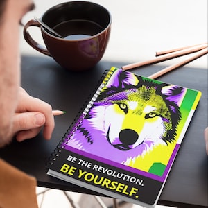 以下が含まれることがあります： 表紙にカラフルなオオカミのイラストが描かれた、スパイラルバインディングのノート。表紙の下部に「BE THE REVOLUTION. BE YOURSELF.」というテキストが印刷されています。