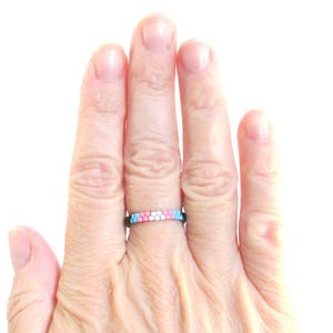 May include: A hand wearing a ring with a pink, blue, and white beaded design.