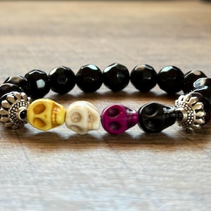 以下が含まれることがあります： 黒色のファセットビーズと、黄色、白、紫、黒の頭蓋骨ビーズが並んだビーズブレスレット。ブレスレットには銀色の装飾的なエンドキャップが付いています。頭蓋骨はユニークなデザイン要素です。