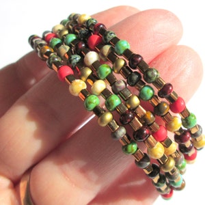 May include: A set of six beaded bracelets with a mix of gold, red, green, brown, and white beads. The bracelets are stacked on top of each other.