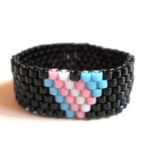 以下が含まれることがあります： 黒ビーズのリングで、トランスジェンダーのプライドフラッグの色、水色、ピンク、白でハート型のデザインが施されています。