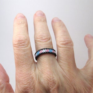 May include: A beaded ring with a black band and a pattern of pink, white, and blue beads.