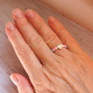 May include: A beaded ring featuring a silver heart charm. The ring is composed of small, colorful beads in shades of pink, blue, and white. The ring is worn on a finger.