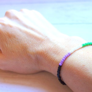 以下が含まれることがあります： 黒、紫、白、緑のビーズで作られたブレスレット。ブレスレットは人の手首につけられています。