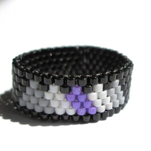 May include: A beaded ring with a black, white, and purple design. The ring is made of small, round beads that are woven together to create a pattern.