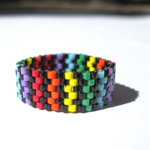 May include: A beaded ring featuring a rainbow color pattern with black accents. The ring showcases a vibrant array of colors, including red, orange, yellow, green, blue, and purple, in a repeating pattern. The ring is approximately 0.75 inches wide.