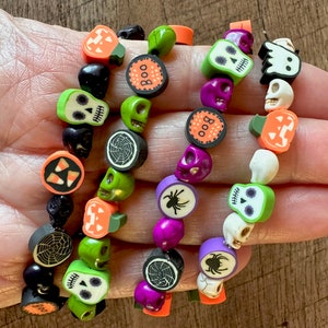 Op de afbeelding: Drie Halloween-armbanden met verschillende bedels, waaronder schedels, pompoenen, snoepmais, spinnen en het woord "Boo".