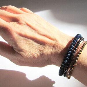 May include: A set of three bracelets, one blue, one black, and one gold, with a beaded design. The bracelets are stacked on a person's wrist.