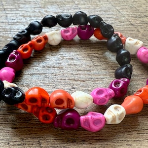 May include: Two bracelets made of skull-shaped beads in various colors, including black, orange, white, pink, and purple. The beads are strung together to form a circular shape. The bracelets are displayed on a wooden surface.