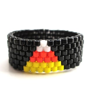 May include: Black beaded ring with a colourful design. The design features a white, red and yellow pattern.