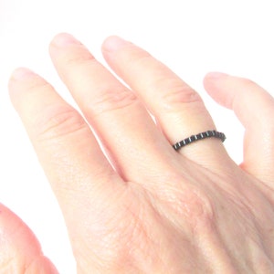 Puede incluir: Un anillo negro con un diseño cuadrado repetitivo. El anillo se lleva en un dedo, mostrando su estilo minimalista. El color oscuro del anillo contrasta con el tono de la piel, resaltando el diseño de la joya. Este anillo es un accesorio simple pero elegante.