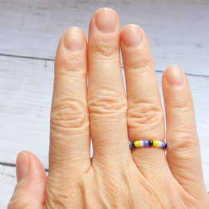 以下が含まれることがあります： 紫、黒、黄色、白のビーズ模様が特徴のビーズリング。指にはめて、カラフルなデザインをアピール。リングはジュエリーです。