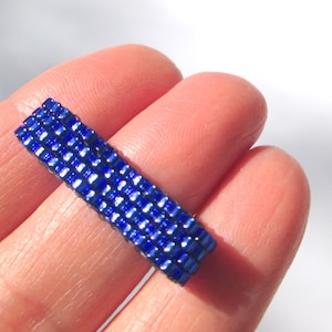 May include: A close-up of a blue beaded bracelet, made of small square beads, on a person's finger.