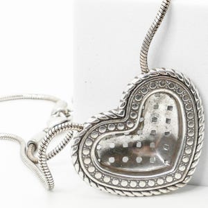 May include: Silver-toned heart-shaped pendant necklace. The pendant has a textured surface with a dotted border and a braided rope design. The necklace chain is a snake chain style.