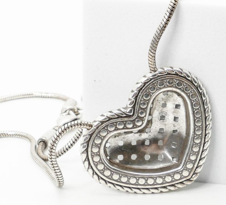 May include: Silver-toned heart-shaped pendant necklace. The pendant has a textured surface with a dotted border and a braided rope design. The necklace chain is a snake chain style.