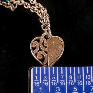 Puede incluir: Un colgante de coraz&oacute;n en tono plateado con un dise&ntilde;o dividido, que muestra un patr&oacute;n de volutas caladas en un lado y una superficie texturizada s&oacute;lida en el otro. El colgante cuelga de una cadena de metal y se mide con una regla de 1,9 cm.