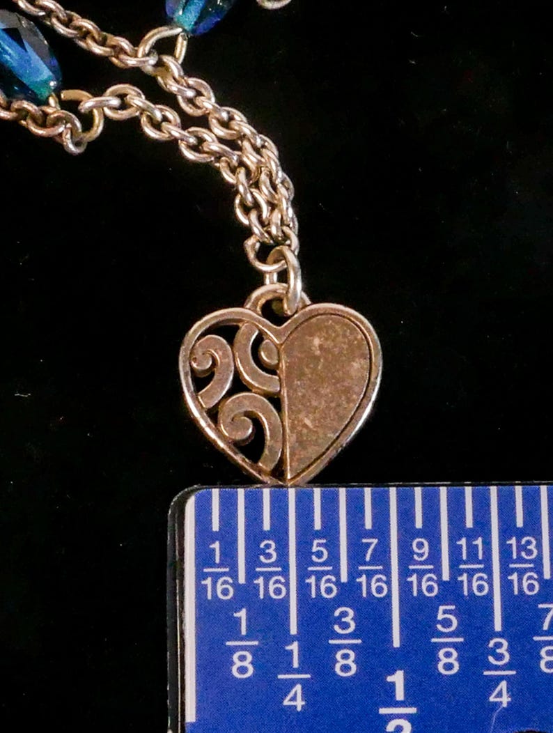 Puede incluir: Un colgante de coraz&oacute;n en tono plateado con un dise&ntilde;o dividido, que muestra un patr&oacute;n de volutas caladas en un lado y una superficie texturizada s&oacute;lida en el otro. El colgante cuelga de una cadena de metal y se mide con una regla de 1,9 cm.
