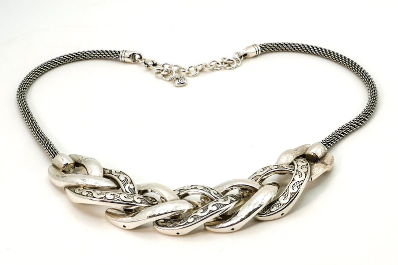 May include: Silver-toned necklace featuring a braided chain design. The necklace has a woven chain and a series of large, textured links with etched detailing. The clasp is a small lobster claw style.