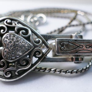 Puede incluir: Un collar con colgante en forma de coraz&oacute;n plateado con un dise&ntilde;o calado y una secci&oacute;n central en forma de coraz&oacute;n incrustada con peque&ntilde;os cristales transparentes. El collar tiene un cierre decorativo y una cadena delicada.