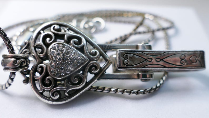 Puede incluir: Un collar con colgante en forma de coraz&oacute;n plateado con un dise&ntilde;o calado y una secci&oacute;n central en forma de coraz&oacute;n incrustada con peque&ntilde;os cristales transparentes. El collar tiene un cierre decorativo y una cadena delicada.