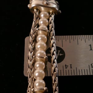 May include: Close-up of a silver-toned necklace pendant with a decorative top. The pendant features a strand of faux pearls and braided silver chains. The ruler shows the pendant is approximately 3 inches in length.