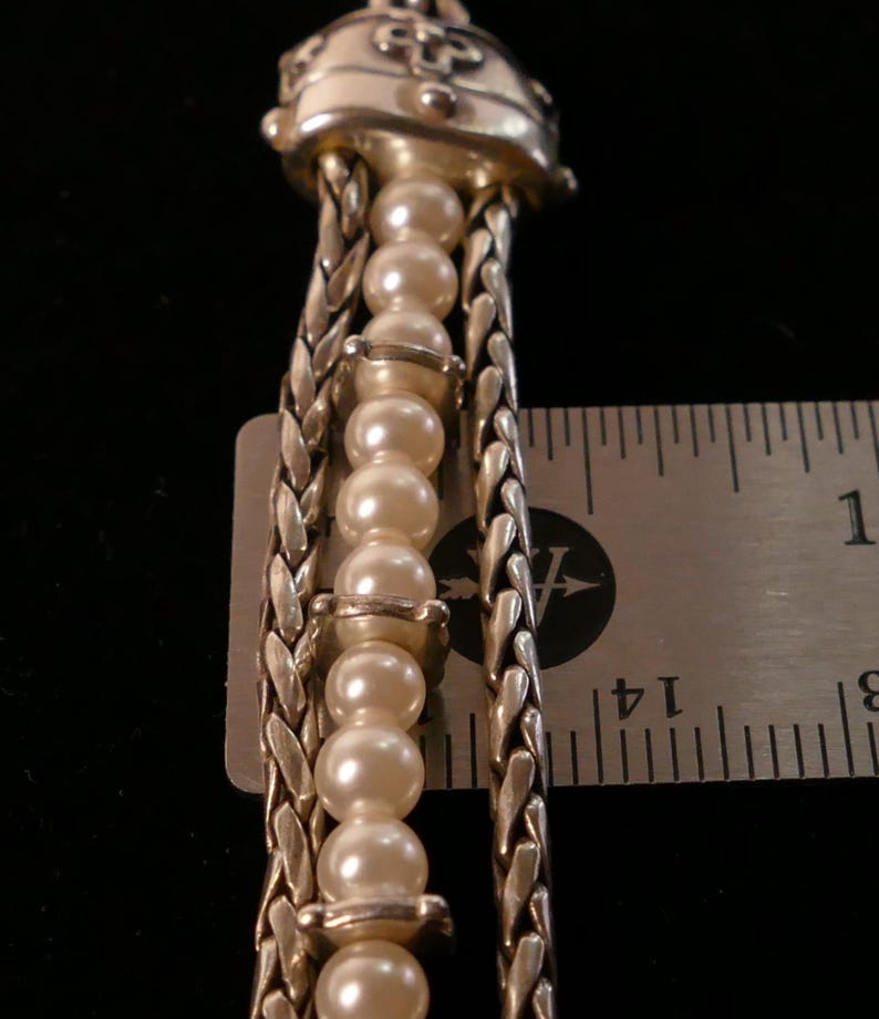 May include: Close-up of a silver-toned necklace pendant with a decorative top. The pendant features a strand of faux pearls and braided silver chains. The ruler shows the pendant is approximately 3 inches in length.