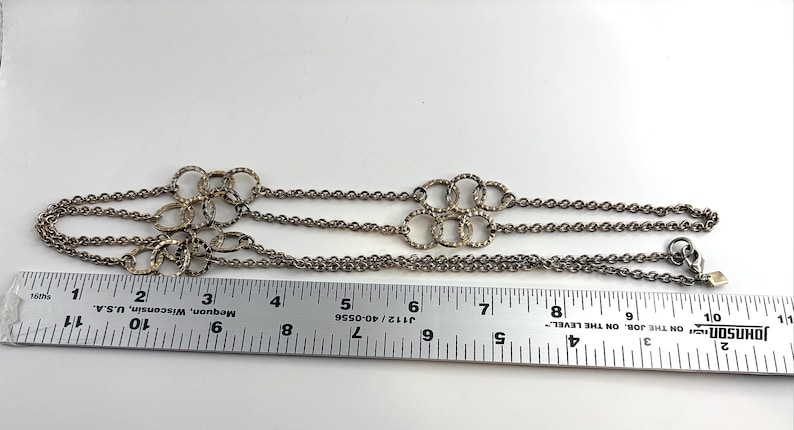 Puede incluir: Collar plateado con cadena y m&uacute;ltiples conjuntos de c&iacute;rculos entrelazados. El collar mide aproximadamente 28 cm de largo, medido con una regla.