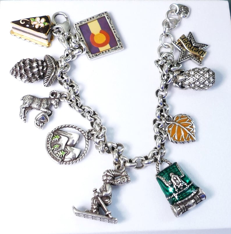 May include: A silver charm bracelet featuring various charms, including a slice of cake, a Colorado flag, a pinecone, a mountain goat, a leaf, and a skier. The charm bracelet is a travel-themed accessory.