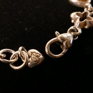 May include: Close-up of a silver bracelet with a heart-shaped clasp and decorative links. The bracelet is on a black background. The clasp is open, showing the connection point.